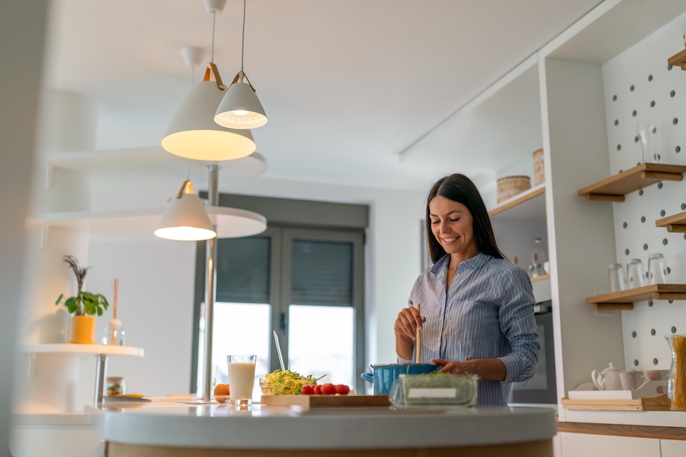 uśmiechnięta kobieta w jasnej kuchni z oświetleniem górnym  kobieta w oświetlonej kuchni mieszająca w garnku